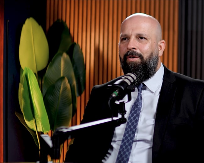 A man with a bald head and a beard sits at a microphone in front of a green plant and wooden paneling.