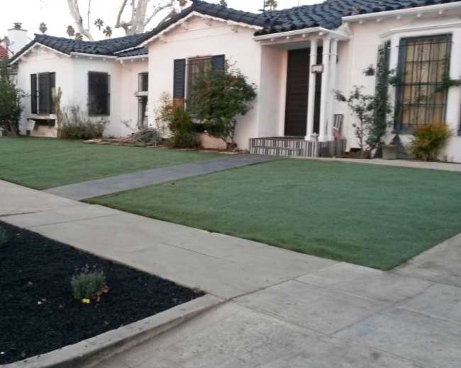 The image shows a residential area with two houses facing a neatly trimmed lawn and pathway, surrounded by low shrubs and landscaping.