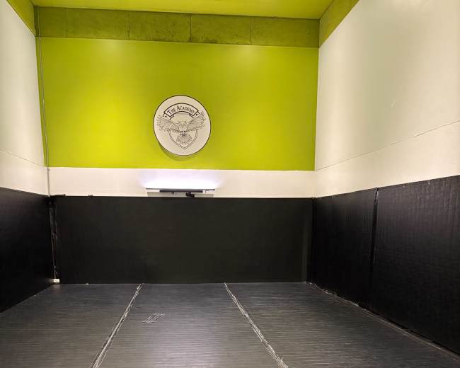 The image shows a spacious, empty training room featuring a black floor mat and a circular logo on the wall with a green accent above it.