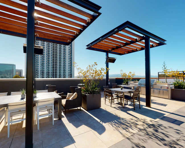 The image shows a rooftop terrace with wooden pergolas, outdoor seating areas, and city buildings in the background.