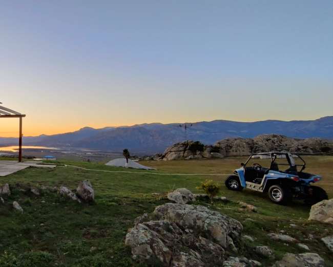 A wooden gazebo stands on a grassy area overlooking a sunset with mountains in the background.