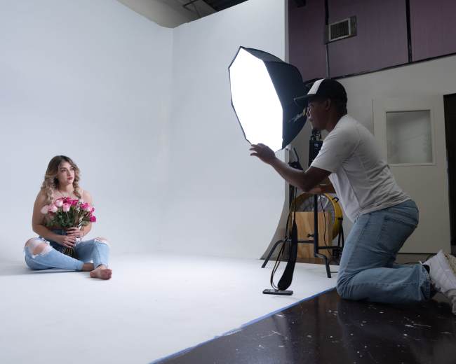 A woman sits on a studio floor holding a bouquet of flowers while a photographer kneels nearby, adjusting a light softbox.