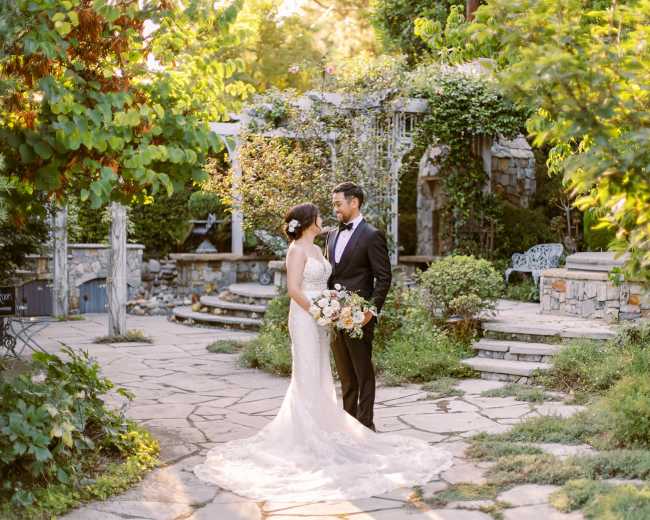 A bride and groom stand together in a stone-walled garden surrounded by greenery and floral arrangements.