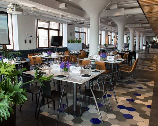 A brightly lit restaurant features neatly arranged tables adorned with flowers and tableware, set against a backdrop of large windows and modern decor.