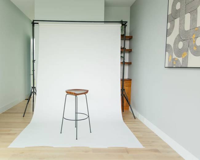 A minimalistic studio setup with a single wooden stool in front of a white backdrop and light-colored wooden flooring.