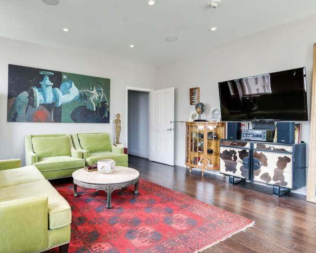 The image shows a modern living room featuring two green sofas, a round coffee table, a red area rug, a large artwork on the wall, and a television mounted on a cabinet.