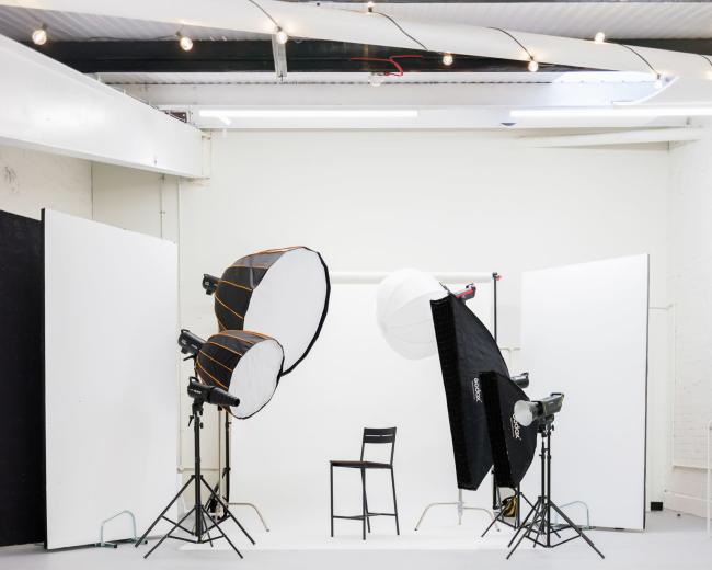 A photography studio is set up with two lighting setups and a single chair positioned in the center against a white backdrop.