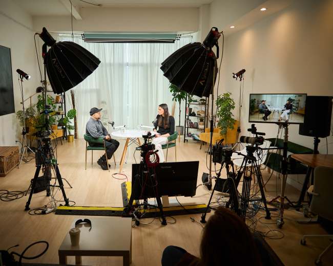 A studio setup features two people seated, surrounded by multiple cameras, lights, and plants, with a large screen displaying content.