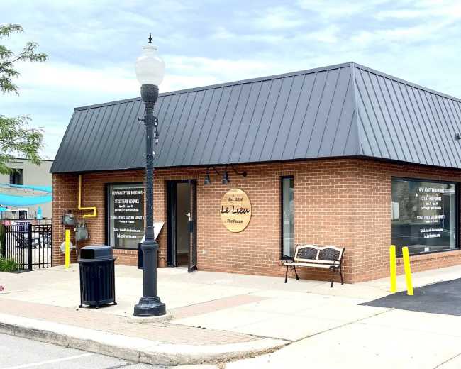 The image shows a single-story brick building with a modern metal roof and large windows displaying business signage, situated on a paved lot with a lamp post and bench nearby.