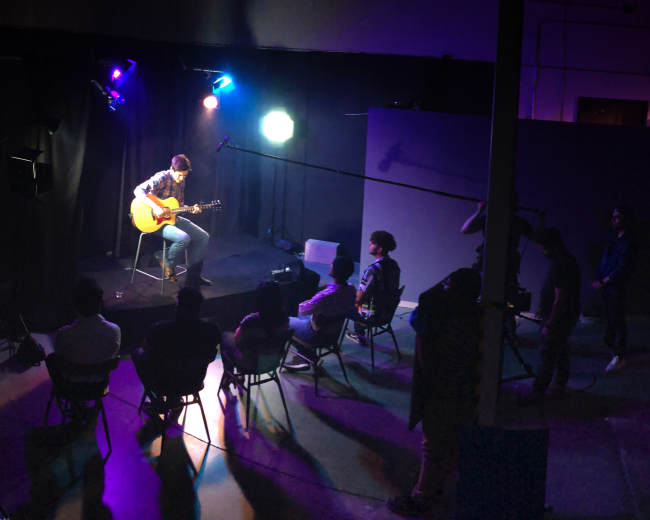 A musician plays guitar on a small stage in a dimly lit room, while an audience sits in chairs facing him.