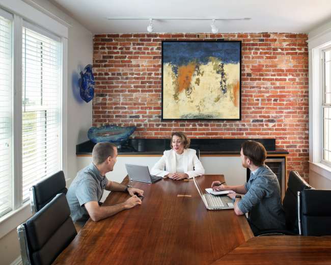 Three people are seated around a wooden conference table in a well-lit room with exposed brick walls and a large painting.