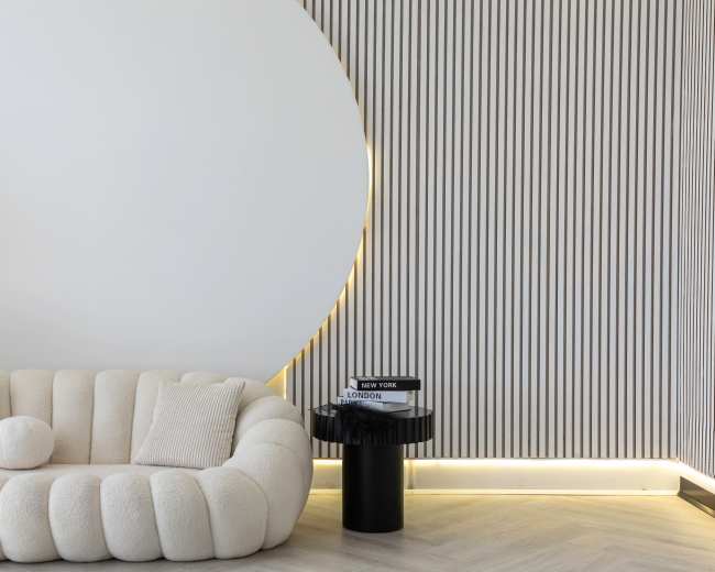 The image features a textured, round white sofa positioned next to a vertical striped wall with ambient lighting and a black circular side table displaying magazines.