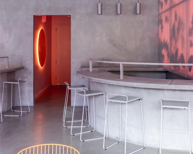 The image shows a modern, minimalist café interior featuring a curved counter, several high stools, and a doorway illuminated with a circular light.