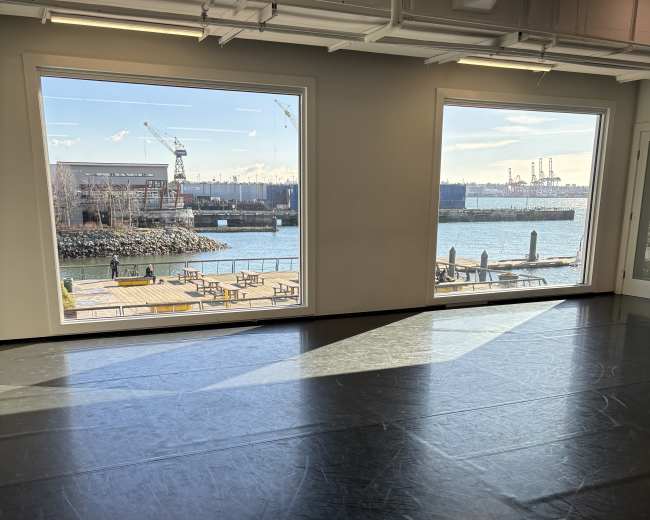 The image shows an indoor space with large windows that overlook a body of water and a dock area, with benches and people visible outside.