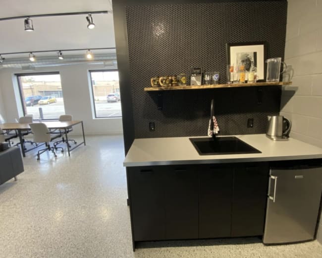 The image shows a modern kitchenette with a black countertop, a sink, a shelf displaying various items, and a small refrigerator, adjacent to a meeting area with tables and chairs.