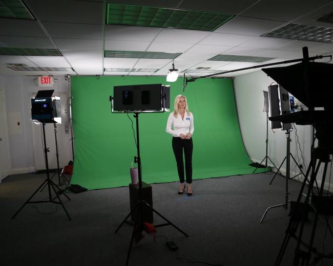 A woman stands in front of a green screen in a studio, surrounded by cameras and lighting equipment.