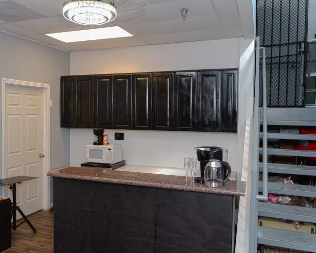 The image shows a small kitchen area with dark cabinets, a countertop, a microwave, a coffee maker, and a staircase in the background.
