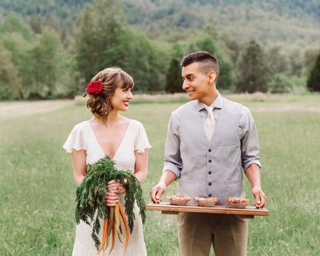 A bride in a white dress holds carrots while a groom in a gray vest presents a wooden tray of desserts in a green field.
