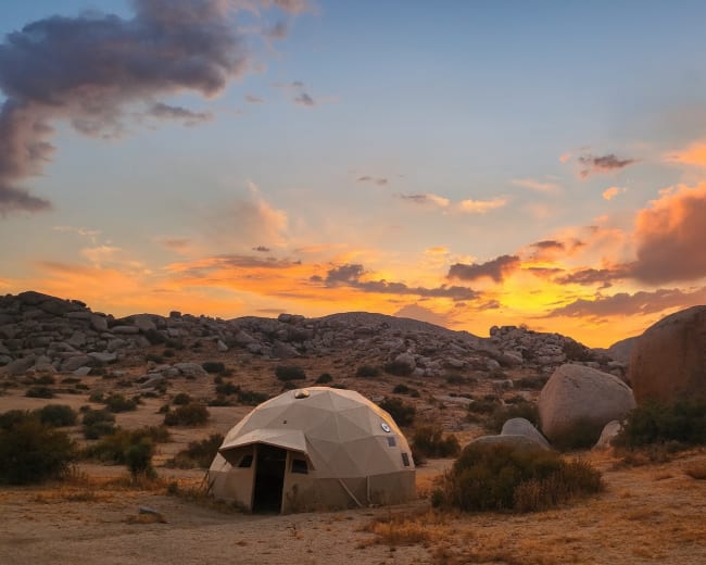 A geodesic dome sits in a rocky desert landscape under a colorful sunset sky.
