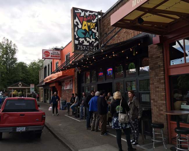 The image shows a group of people gathered outside a café with a colorful sign, while a red truck is parked nearby on a street.