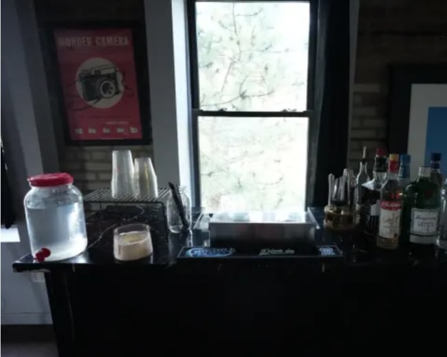 The image shows a bar setup with a variety of liquor bottles, a glass of drink, cups, and a large jar of liquid on a dark countertop next to a window.