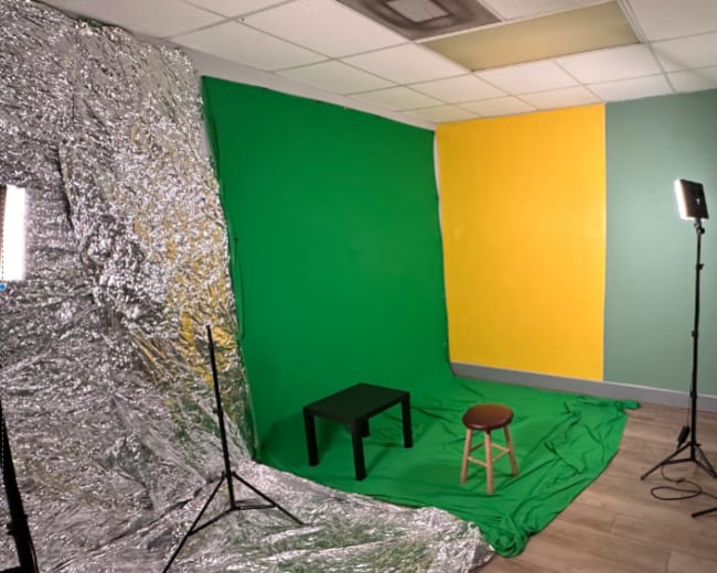 A small studio setup with green and yellow backdrops, a table, a stool, and lighting equipment.