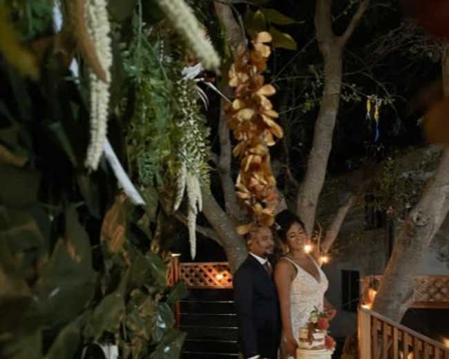 A couple stands beside a wedding cake on a table decorated with flowers in a softly lit outdoor venue surrounded by trees.