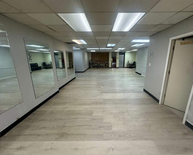 The image shows an empty room with wooden flooring, bright overhead lights, and mirrors on one wall.
