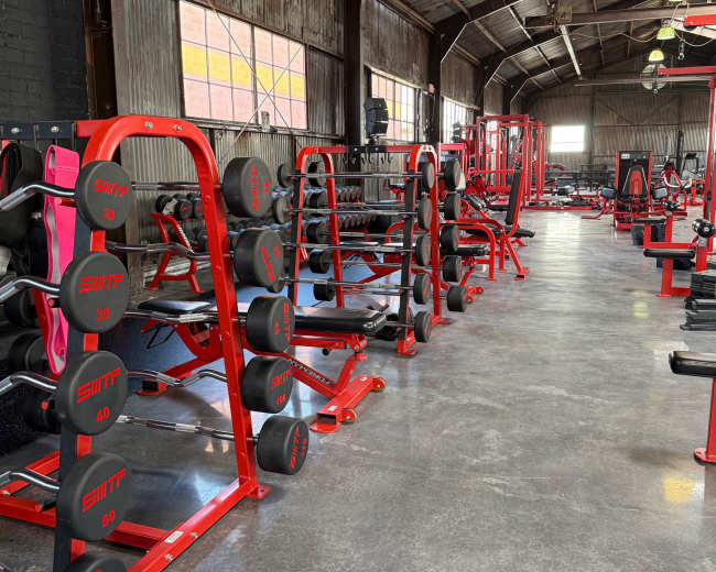 The image shows a spacious gym with red weightlifting equipment and organized rows of dumbbells on racks.
