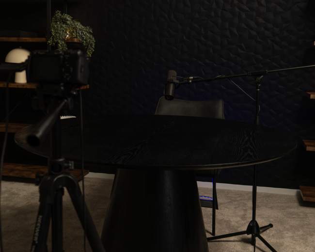 A black table with a conical base is set up in a dimly lit room, accompanied by a chair, a microphone, and a camera on a tripod.