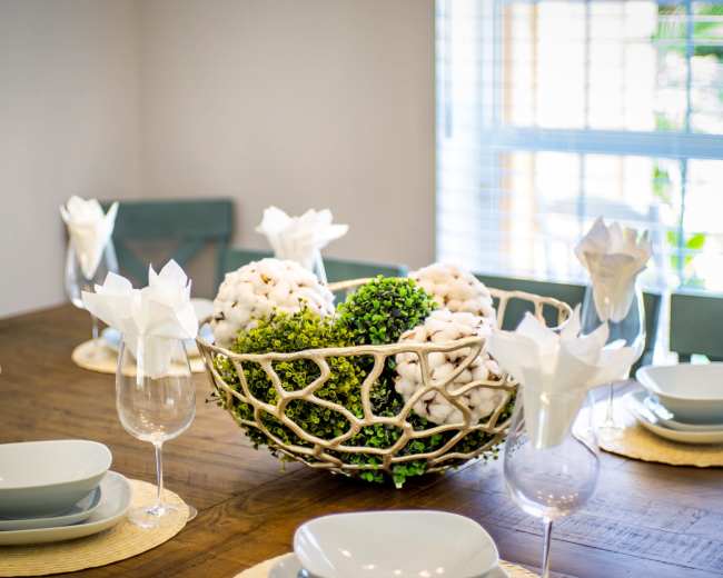 A dining table is set with grey plates and glasses, featuring a decorative bowl of cotton and greenery at the center.