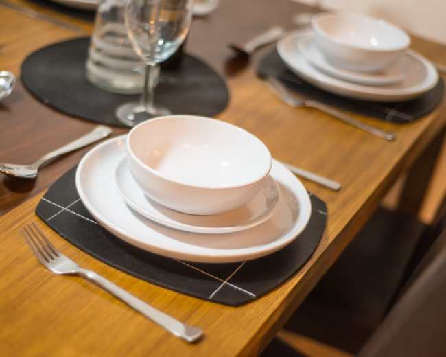 A dining table is set with white plates, bowls, and silverware on black placemats.