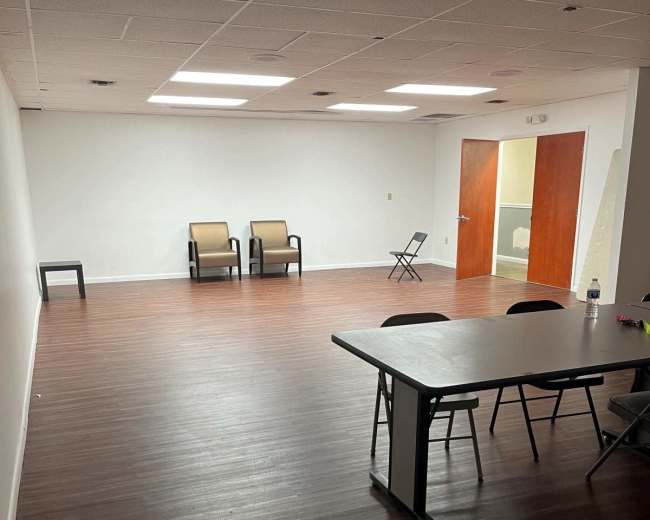 The image shows a spacious, empty room with brown wooden floors, two chairs against one wall, and a table with chairs set up in the foreground.