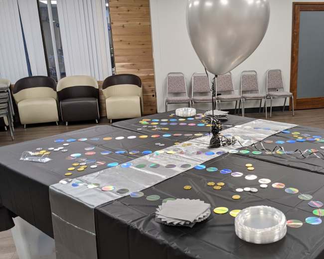 The image shows a decorated table in a room with folding chairs, featuring colorful circular decorations and a silver balloon.