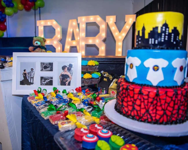 A colorful baby-themed celebration features a decorated cake, treats, and a photo display, with large illuminated letters spelling "BABY" in the background.