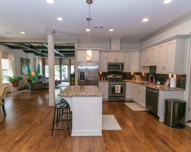 The image shows a modern kitchen with a central island, stainless steel appliances, and wooden flooring, connected to a living area with natural light.
