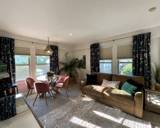 A cozy living room features a brown sofa, a glass dining table with pink chairs, and large windows covered with floral curtains.