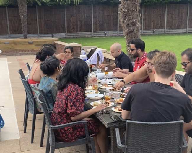 A group of people sits around a large outdoor table, sharing a meal in a backyard setting.