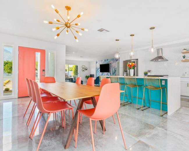 The image depicts a modern kitchen and dining area with a wooden table surrounded by coral-colored chairs and a turquoise kitchen island.