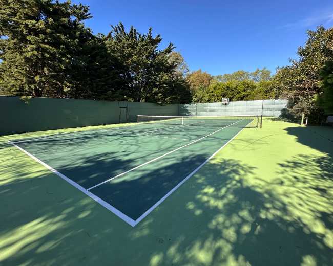 The image shows a vacant tennis court surrounded by trees and a clear blue sky.