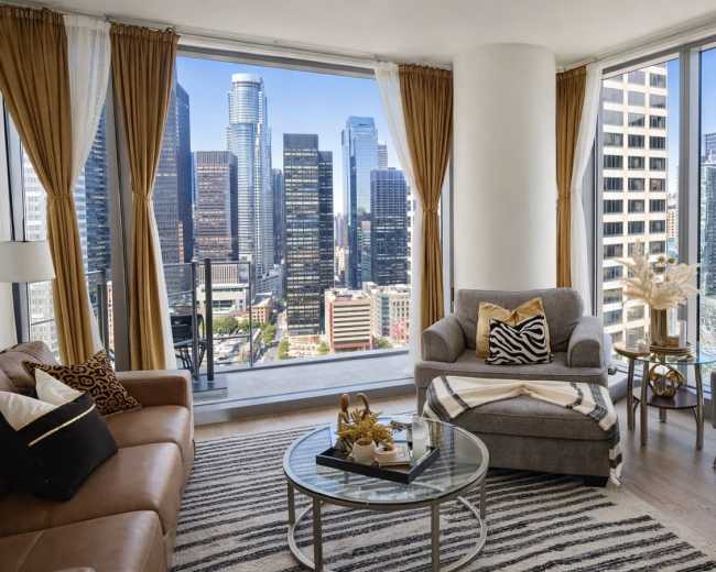 The image shows a modern apartment living room with large windows offering a view of a city skyline, featuring a mix of seating arrangements and decorative elements.