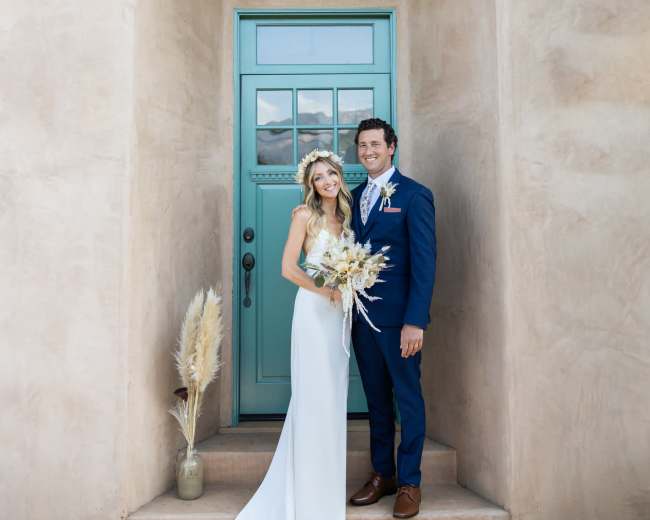 A bride and groom pose together at the entrance of a house with a teal door, holding a bouquet of dried flowers.