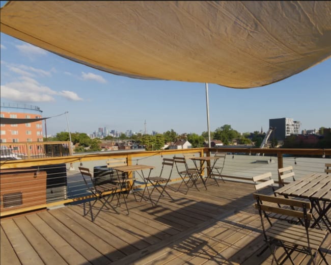 A rooftop terrace features wooden tables and chairs under a large canopy, with a city skyline visible in the background.