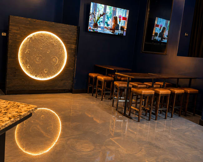The image shows a modern restaurant interior featuring a dark blue wall with a circular wall design, a television mounted above, and wooden tables with stools arranged in front.