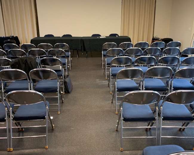 A meeting room with rows of blue chairs facing a table at the front against a light-colored wall.