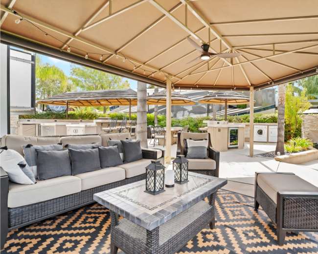 A shaded outdoor seating area features a stylish sofa, coffee table, and lanterns, surrounded by cooking stations and greenery.