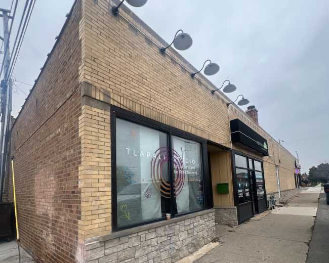 The image shows a corner commercial building with a brick exterior, large windows featuring colorful designs, and a sidewalk in front.