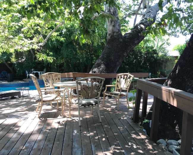 A wooden deck features white outdoor furniture surrounded by trees, with a swimming pool visible in the background.