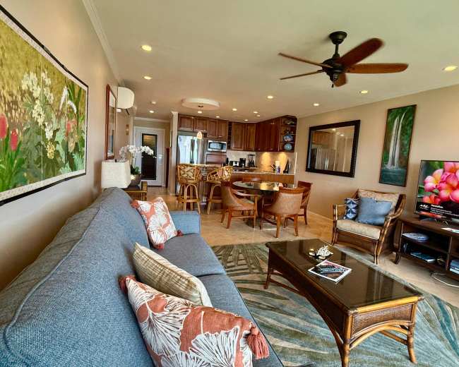 The image shows a well-furnished living area with a sofa, coffee table, and dining space, featuring modern appliances in an open kitchen.