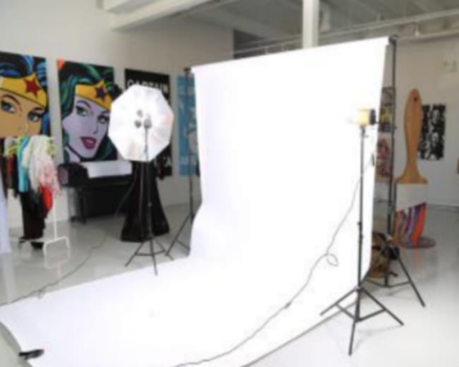 The image shows a photography studio setup with a white backdrop, studio lights, and various clothing items in the background.